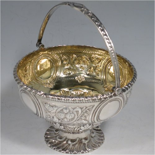 An Antique Victorian Sterling Silver sweet-meat basket, having a round baluster body with gold-gilt interior and an applied gadroon border, with hand-chased floral decoration between four round vacant cartouches, a hinged and florally chased swing handle, and all sitting on a swirl-fluted pedestal foot. Made by Martin and Hall of London in 1871. The dimensions of this fine hand-made antique silver basket are height 15 cms (6 inches), diameter 12.5 cms (5 inches), and it weighs approx. 224g (7.2 troy ounces).