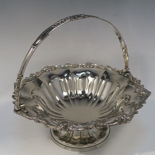 A very pretty Antique Victorian Silver-Plated basket, having a round body with hand-chased fluted decoration and an applied shell and scroll border, a hinged swing handle with applied floral decoration, and all sitting on a round fluted pedestal foot. This beautiful silver-plated swing-handle basket was made by Walker and Hall of Sheffield in ca. 1880. The dimensions of this fine hand-made antique silver-plated basket are height (inc. handles) 25.5 cms (10 inches), and diameter  28 cms (11 inches),   