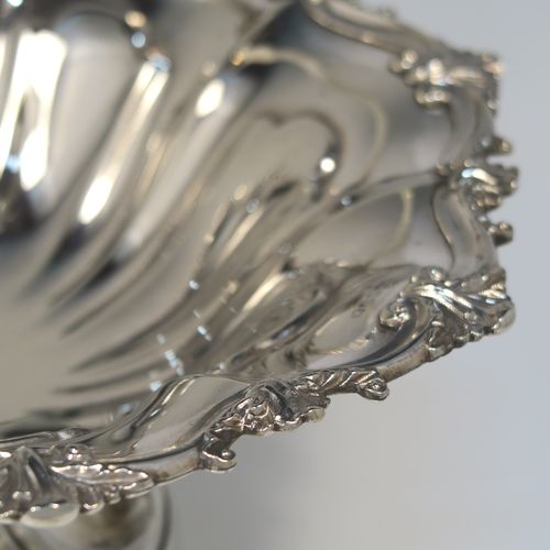 A very pretty Antique Victorian Silver-Plated basket, having a round body with hand-chased fluted decoration and an applied shell and scroll border, a hinged swing handle with applied floral decoration, and all sitting on a round fluted pedestal foot. This beautiful silver-plated swing-handle basket was made by Walker and Hall of Sheffield in ca. 1880. The dimensions of this fine hand-made antique silver-plated basket are height (inc. handles) 25.5 cms (10 inches), and diameter  28 cms (11 inches),   
