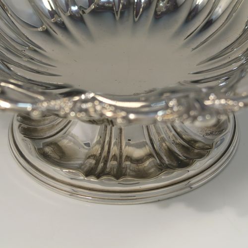 A very pretty Antique Victorian Silver-Plated basket, having a round body with hand-chased fluted decoration and an applied shell and scroll border, a hinged swing handle with applied floral decoration, and all sitting on a round fluted pedestal foot. This beautiful silver-plated swing-handle basket was made by Walker and Hall of Sheffield in ca. 1880. The dimensions of this fine hand-made antique silver-plated basket are height (inc. handles) 25.5 cms (10 inches), and diameter  28 cms (11 inches),   