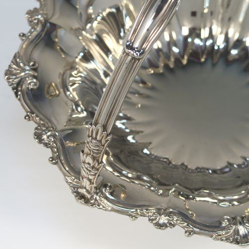 A very pretty Antique Victorian Silver-Plated basket, having a round body with hand-chased fluted decoration and an applied shell and scroll border, a hinged swing handle with applied floral decoration, and all sitting on a round fluted pedestal foot. This beautiful silver-plated swing-handle basket was made by Walker and Hall of Sheffield in ca. 1880. The dimensions of this fine hand-made antique silver-plated basket are height (inc. handles) 25.5 cms (10 inches), and diameter  28 cms (11 inches),   