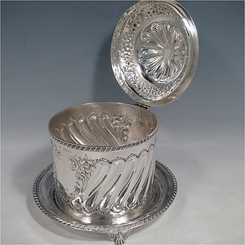 An Antique Victorian Sterling Silver biscuit box, having a round straight-sided body with hand-chased fluting and floral decoration, a hinged lid with cast foliate finial, and sitting on an attached base plate with a gadroon border and three cast foliate & lions-paw feet. Made by William Hutton & Sons of London in 1899. The dimensions of this fine hand-made antique silver biscuit box are height 18 cms (7 inches), diameter at base 19 cms (7.5 inches), and it weighs approx. 744g (24 troy ounces).