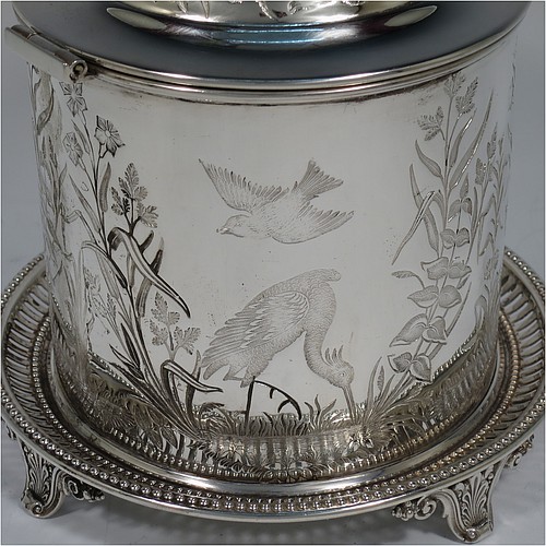 An Antique Victorian Silver Plated Chinoiserie style biscuit box, having a round body with straight sides and applied bead-edged borders, with very pretty hand-engraved flowers, reeds, birds, and butterfly work, a hinged domed lid with cast finial, and all sitting on a hand-pierced attached plate with four cast scroll feet. Made by the Atkin Brothers of Sheffield in ca. 1880. The dimensions of this fine hand-made antique silver-plated biscuit box are height 18 cms (7 inches), and diameter at base 17.5 cms (7 inches).    