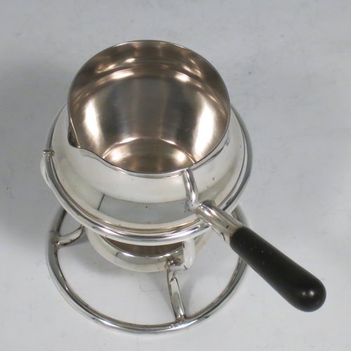 A Sterling Silver brandy saucepan warmer on burner stand, having a plain round baluster body, a sparrow-beak spout, an original wood turned handle, and sitting on a wire-work frame with integral spirit burner. Made by Hukin & Heath of Birmingham in 1936. The dimensions of this fine hand-made silver brandy warmer are height 8 cms (3.25 inches), length (inc. handle) 12 cms (4.75 inches), and it weighs approx. 105g (3.4 troy ounces).