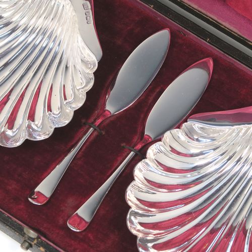 An elegant Antique Victorian Sterling Silver pair of butter shells, having traditional scallop shapes, and sitting on three cast ball feet, together with a pair of original butter knives in the Old English pattern, and all sitting in an original maroon satin and velvet-lined presentation box. This handsome pair of silver butter shells and knives were all made by the Atkin Brothers of Sheffield in 1900. The dimensions of this fine hand-made set of antique silver butter shells and knives are length 12 cms (4.75 inches), width 10 cms (4 inches), with a total weight of approx. 136g (4.4 troy ounces).