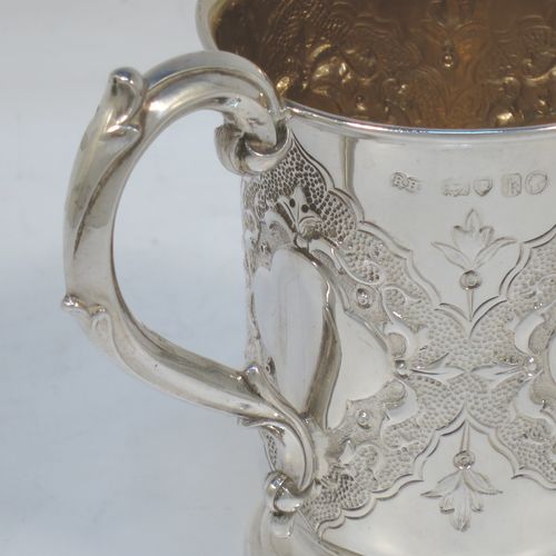 A very pretty Antique Victorian Sterling Silver christening mug, having a round body with straight tapering sides and tucked under belly, with hand-chased floral and scroll-work decoration surrounding four vacant lobed cartouches, a scroll loop handle, a gold-gilt interior, and all sitting on a pedestal foot. Made by Robert Hennell of London in 1865. The dimensions of this fine hand-made antique silver christening mug are height 10 cms (4 inches), diameter at lip 7 cms (2.75 inches), and it weighs approx. 130g (4.2 troy ounces).  