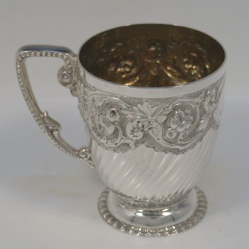 A very pretty Antique Victorian Sterling Silver christening mug, having a round tapering sides, all hand-chased with floral, scroll-work, and fluted decoration, a cast and bead-edged scroll handle, a central vacant cartouche opposite the handle, a gold-gilt interior, and all sitting on a pedestal foot. This beautiful silver christening mug was made by Wakely and Wheeler of London in 1901. The dimensions of this fine hand-made antique silver christening mug are height 9.5 cms (3.75 inches), diameter at lip 7.5 cms (3 inches), and it weighs approx. 161g (5.2 troy ounces).  