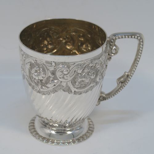 A very pretty Antique Victorian Sterling Silver christening mug, having a round tapering sides, all hand-chased with floral, scroll-work, and fluted decoration, a cast and bead-edged scroll handle, a central vacant cartouche opposite the handle, a gold-gilt interior, and all sitting on a pedestal foot. This beautiful silver christening mug was made by Wakely and Wheeler of London in 1901. The dimensions of this fine hand-made antique silver christening mug are height 9.5 cms (3.75 inches), diameter at lip 7.5 cms (3 inches), and it weighs approx. 161g (5.2 troy ounces).  