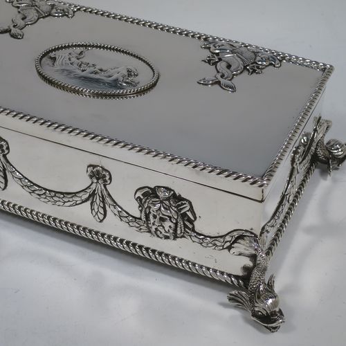 A very handsome and unusual Neptune themed Antique Victorian Sterling Silver cigar / cigarette box, having a rectangular body with applied rope-twist borders, laurel-leaf swags, Neptune face masks, and inter-twined dolphins, a hinged lid with an applied central oval cartouche with a scene of Neptune, mermaids, and sea-horses, a varnished wood-lined interior, and all sitting on four cast dolphin feet. Made by Reid and Sons of London in 1901. The dimensions of this fine hand-made antique silver table box are length 26 cms (10 inches), width 14 cms (5.5 inches), and height 8 cms (3 inches).