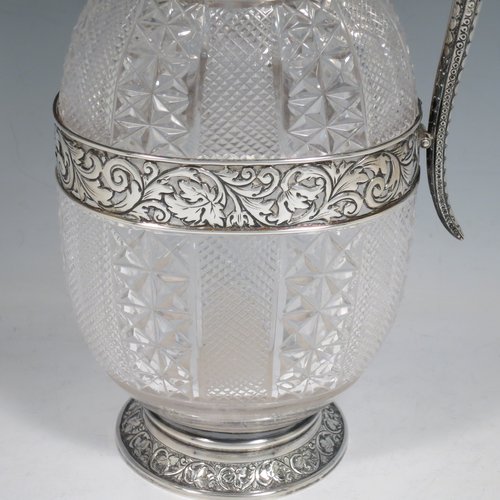 An Antique Victorian Sterling Silver and hand-cut crystal claret jug, having a hand-chased mount with floral decoration and a wavey bead-edged border, a hinged lid with cast floral finial, a hand-chased and engraved scroll handle attached to a hand-pierced floral band, together with an oval cross-section hand-cut Webb crystal body with star-cut panels, all sitting on a matching collet foot. Made by Gibson & Langland of London in 1890. The dimensions of this fine hand-made antique silver and crystal claret jug are height 31 cms (12.25 inches), and length 15 cms (6 inches).
