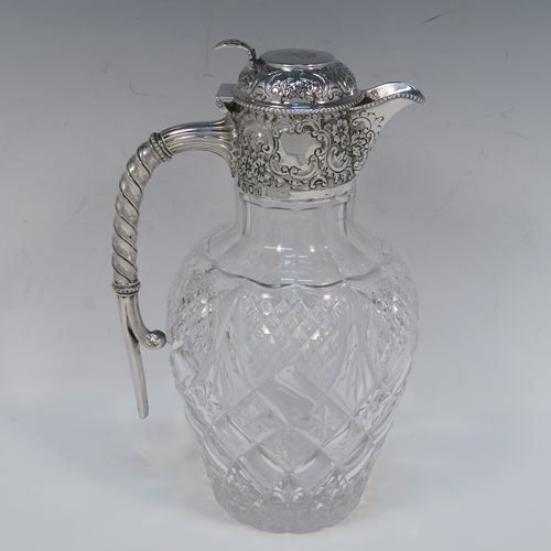 A very beautiful Antique Victorian Sterling Silver and hand-cut crystal claret jug, having an ornate round mount with hand-chased floral and scroll decoration, a fluted and twisted style side handle, a hinged lid with matching decoration and a pierced thumb-piece, together with a hand-cut crystal round body with tapering sides and above a flat star-cut base. This beautiful antique silver and crystal claret jug was made by Gibson and Langland of London in 1898. The dimensions of this fine hand-made antique silver and crystal claret jug are height 23 cms (9 inches), and length 14 cms (5.5 inches). Please note that this item is crested on the top of the lid.