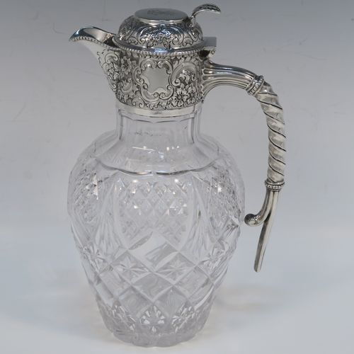 A very beautiful Antique Victorian Sterling Silver and hand-cut crystal claret jug, having an ornate round mount with hand-chased floral and scroll decoration, a fluted and twisted style side handle, a hinged lid with matching decoration and a pierced thumb-piece, together with a hand-cut crystal round body with tapering sides and above a flat star-cut base. This beautiful antique silver and crystal claret jug was made by Gibson and Langland of London in 1898. The dimensions of this fine hand-made antique silver and crystal claret jug are height 23 cms (9 inches), and length 14 cms (5.5 inches). Please note that this item is crested on the top of the lid.