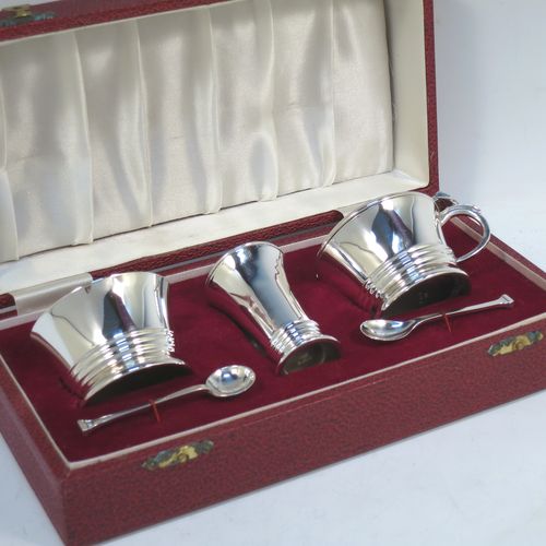A very elegant Sterling Silver three-piece condiment set in an Art Deco style, consisting of an open salt cellar with a blue glass liner, a pepper pot with pull-off lid, a mustard pot with blue glass liner and hinged lid, together with two original condiment spoons, and all in their cream satin and maroon velvet-lined presentation box. Made by Deakin and Francis of Birmingham in 1970. The dimensions of this fine hand-made silver condiment service are length of mustard pot 7.5 cms (3 inches), height of pepper pot 7 cms (2.75 inches), diameter of salt cellar 6 cms (2.3 inches), and with a total weight of approx. 164g (5.3 troy ounces). 