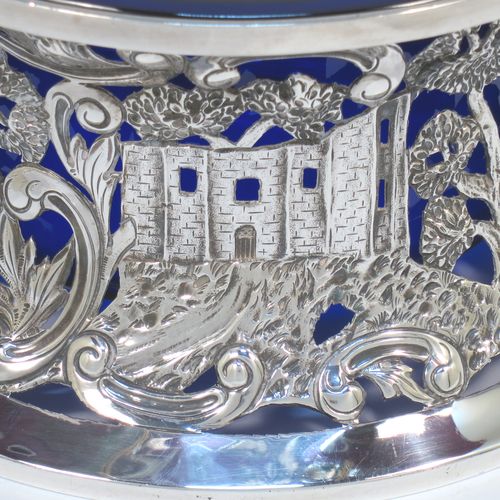 A very beautiful Irish Antique Edwardian Sterling Silver large table dish ring, having a hand-pierced and chased body decorated with scrolls, two cottages, a castle, and a bridge, a vacant oval cartouche, and a blue-glass liner. Made by Edmund Johnson of Dublin in 1910. The dimensions of this fine hand-made antique silver dish ring are height 9 cms (3.5 inches), diameter at base 19 cms (7.5 inches), and it weighs approx. 302g (9.7 troy ounces).