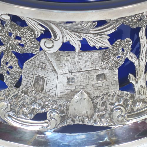 A very beautiful Irish Antique Edwardian Sterling Silver large table dish ring, having a hand-pierced and chased body decorated with scrolls, two cottages, a castle, and a bridge, a vacant oval cartouche, and a blue-glass liner. Made by Edmund Johnson of Dublin in 1910. The dimensions of this fine hand-made antique silver dish ring are height 9 cms (3.5 inches), diameter at base 19 cms (7.5 inches), and it weighs approx. 302g (9.7 troy ounces).