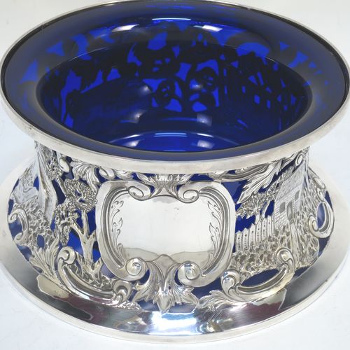 A very beautiful Irish Antique Edwardian Sterling Silver large table dish ring, having a hand-pierced and chased body decorated with scrolls, two cottages, a castle, and a bridge, a vacant oval cartouche, and a blue-glass liner. Made by Edmund Johnson of Dublin in 1910. The dimensions of this fine hand-made antique silver dish ring are height 9 cms (3.5 inches), diameter at base 19 cms (7.5 inches), and it weighs approx. 302g (9.7 troy ounces).