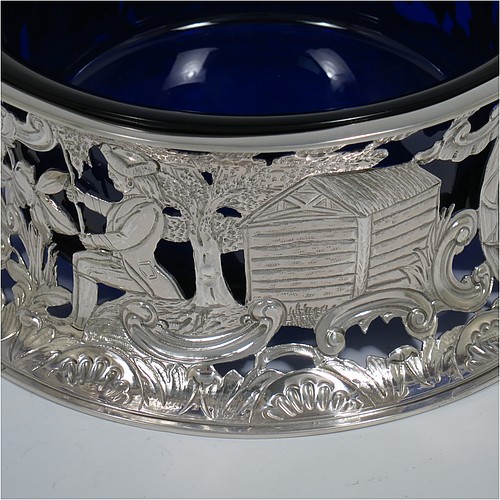 An Antique Victorian Sterling Silver large table dish ring in an Irish style, having a hand-pierced and chased body decorated with pastoral scenes, figures, animals, a vacant oval cartouche, and a blue-glass liner. Made by D. and J. Welby of London in 1894. The dimensions of this fine hand-made antique silver dish ring are height 7.5 cms (3 inches), diameter at base 19 cms (7.5 inches), and it weighs approx. 351g (11.3 troy ounces).