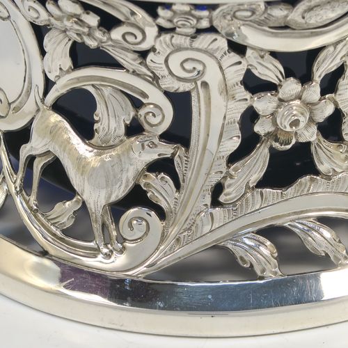 A very beautiful Irish Antique Sterling Silver large table dish ring, having a hand-pierced and chased body decorated with scrolls, flowers, and animals, a single vacant cartouche, and a blue-glass liner. This elegant silver potato or dish ring was made by West and Son of Dublin in 1917. The dimensions of this fine hand-made antique silver dish ring are height 10 cms (4 inches), diameter at base 18.5 cms (7.25 inches), and it weighs approx. 326g (10.5 troy ounces).