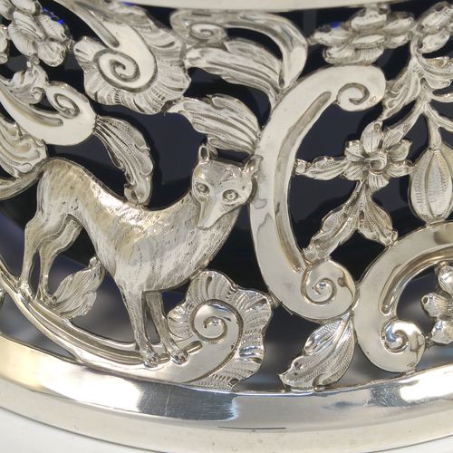 A very beautiful Irish Antique Sterling Silver large table dish ring, having a hand-pierced and chased body decorated with scrolls, flowers, and animals, a single vacant cartouche, and a blue-glass liner. This elegant silver potato or dish ring was made by West and Son of Dublin in 1917. The dimensions of this fine hand-made antique silver dish ring are height 10 cms (4 inches), diameter at base 18.5 cms (7.25 inches), and it weighs approx. 326g (10.5 troy ounces).