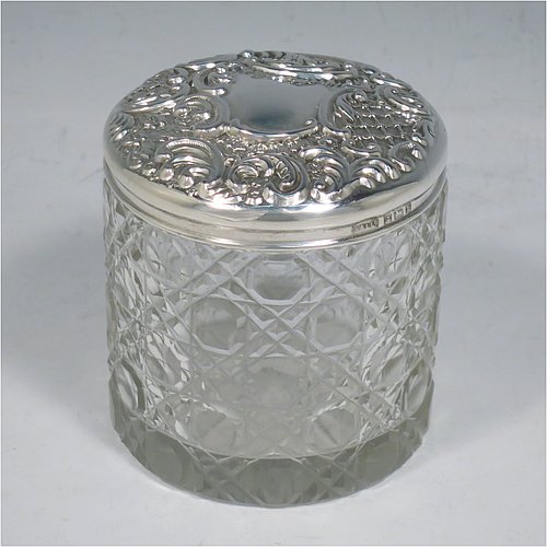 An Antique Victorian Sterling Silver and cut-crystal dressing table jar / box, having a round hand-cut crystal body in the Hobnail pattern, a hand-chased pull-off lid with floral decoration, and sitting on a flat star-cut base. Made by Adie & Lovekin Ltd., of Birmingham in 1901. The dimensions of this fine hand-made antique silver and crystal dressing table box / jar are height 8 cms (3 inches), and diameter 7 cms (2.75 inches).   