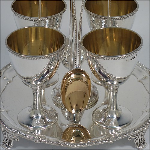 An Antique Victorian Sterling Silver egg cruet set, having four plain round egg cups with gold-gild interiors sitting on pedestal feet, four egg spoons with gold-gilt bowls, and a plain cast loop handle attached to a round base with an applied shaped gadroon border, and sitting on four cast scroll feet. Made by George Hancock of Sheffield in 1897 (the spoons by James Dixon and Sons of Sheffield in 1917). The dimensions of this fine hand-made antique silver egg cruet set are height 20 cms (8 inches), diameter of base is 15 cms (6 inches), and the total weight is approx. 688g (22 troy ounces).