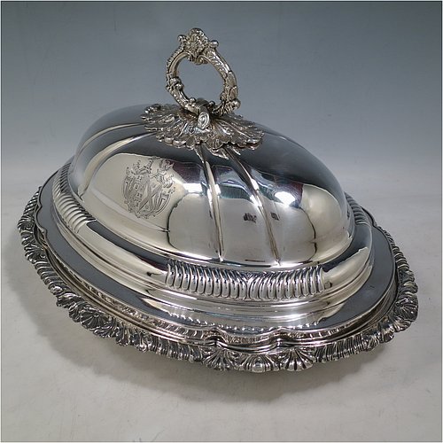 An Antique Georgian pair of Old Sheffield Plated entrée dishes with covers, having shaped oval bodies with with cast shell and gadroon borders, domed lids with hand-chased fluted decoration, and removable cast oak-leaf handles, and all sitting on flat bases. Made in ca. 1820. The dimensions of this pair of fine hand-made antique Georgian Old Sheffield Plated entrée dishes and covers are length 36 cms (14 inches), height including handle 21.5 cms (8.5 inches), and width 27.5 cms (10.75 inches). Please note that these are engraved with full armorial crests.