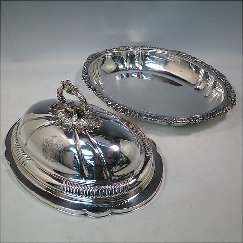 An Antique Georgian pair of Old Sheffield Plated entrée dishes with covers, having shaped oval bodies with with cast shell and gadroon borders, domed lids with hand-chased fluted decoration, and removable cast oak-leaf handles, and all sitting on flat bases. Made in ca. 1820. The dimensions of this pair of fine hand-made antique Georgian Old Sheffield Plated entrée dishes and covers are length 36 cms (14 inches), height including handle 21.5 cms (8.5 inches), and width 27.5 cms (10.75 inches). Please note that these are engraved with full armorial crests.
