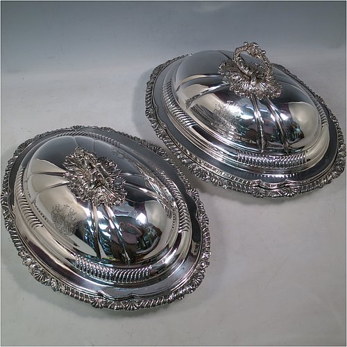 An Antique Georgian pair of Old Sheffield Plated entrée dishes with covers, having shaped oval bodies with with cast shell and gadroon borders, domed lids with hand-chased fluted decoration, and removable cast oak-leaf handles, and all sitting on flat bases. Made in ca. 1820. The dimensions of this pair of fine hand-made antique Georgian Old Sheffield Plated entrée dishes and covers are length 36 cms (14 inches), height including handle 21.5 cms (8.5 inches), and width 27.5 cms (10.75 inches). Please note that these are engraved with full armorial crests.