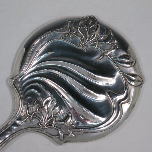 An Antique Edwardian Art Nouveau Sterling Silver hand-mirror, having a round body with hand-chased floral lilly decoration, and an original round bevelled mirror with rope-twist border. Made by Henry Williamson Ltd., of Birmingham in 1905. The dimensions of this fine hand-made antique silver hand mirror are length 28 cms (11 inches), and diameter 15 cms (6 inches). Please note that the mirror glass has very slight scratches concurrent with its age.