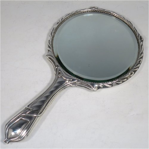 An Antique Edwardian Art Nouveau Sterling Silver hand-mirror, having a round body with hand-chased floral lilly decoration, and an original round bevelled mirror with rope-twist border. Made by Henry Williamson Ltd., of Birmingham in 1905. The dimensions of this fine hand-made antique silver hand mirror are length 28 cms (11 inches), and diameter 15 cms (6 inches). Please note that the mirror glass has very slight scratches concurrent with its age.