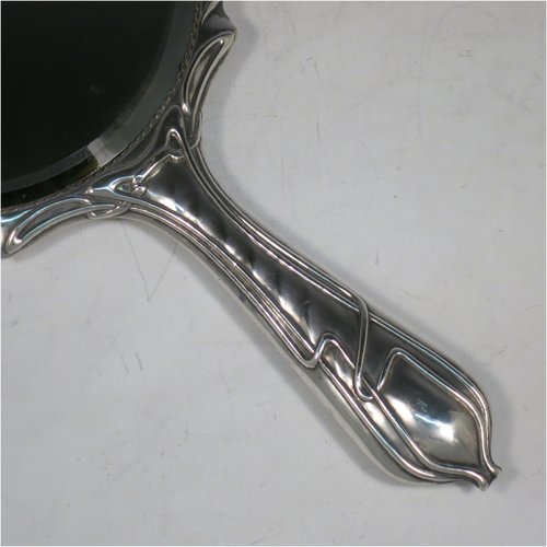 An Antique Edwardian Art Nouveau Sterling Silver hand-mirror, having a round body with hand-chased floral lilly decoration, and an original round bevelled mirror with rope-twist border. Made by Henry Williamson Ltd., of Birmingham in 1905. The dimensions of this fine hand-made antique silver hand mirror are length 28 cms (11 inches), and diameter 15 cms (6 inches). Please note that the mirror glass has very slight scratches concurrent with its age.