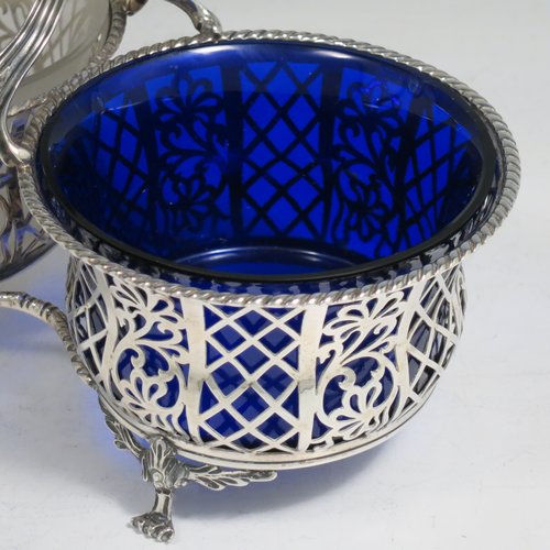 An Antique Edwardian Sterling Silver preserve, jam and honey pot holder, having a blue and frosted glass pot, sitting in a hand-pierced frame with floral and geometric decoration, with applied gadroon borders, a reeded and pierced loop handle, and all sitting on four cast floral feet. Made by Mappin and Webb of London in 1907. The dimensions of this fine hand-made antique silver and glass preserve holder are length 21 cms (8.25 inches), width 11 cms (4.25 inches), height 17.5 cms (7 inches), and it weighs approx. 243g (7.8 troy ounces).