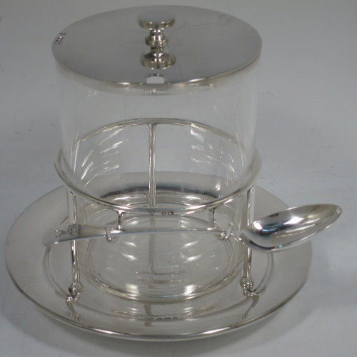 A very unusual, large, and handsome pair of Antique Victorian sterling silver and hand-cut crystal jam, honey, or preserve jars, having round straight-sided bodies with plain pull-off lids with disc finials, and sitting in gallery frames with two hooks for holding jam or honey spoons, and attached to round base plates with three cushion feet underneath. Made by Charles Edward Nixon of Sheffield in 1896. The dimensions of these fine hand-made silver and crystal jam, honey, or conserve jars are height 13.5 cms (5.25 inches), diameter at base 15 cms (6 inches) square. Please note that these come with two matching silver serving spoons.