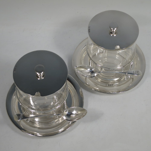 A very unusual, large, and handsome pair of Antique Victorian sterling silver and hand-cut crystal jam, honey, or preserve jars, having round straight-sided bodies with plain pull-off lids with disc finials, and sitting in gallery frames with two hooks for holding jam or honey spoons, and attached to round base plates with three cushion feet underneath. Made by Charles Edward Nixon of Sheffield in 1896. The dimensions of these fine hand-made silver and crystal jam, honey, or conserve jars are height 13.5 cms (5.25 inches), diameter at base 15 cms (6 inches) square. Please note that these come with two matching silver serving spoons.