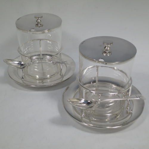 A very unusual, large, and handsome pair of Antique Victorian sterling silver and hand-cut crystal jam, honey, or preserve jars, having round straight-sided bodies with plain pull-off lids with disc finials, and sitting in gallery frames with two hooks for holding jam or honey spoons, and attached to round base plates with three cushion feet underneath. Made by Charles Edward Nixon of Sheffield in 1896. The dimensions of these fine hand-made silver and crystal jam, honey, or conserve jars are height 13.5 cms (5.25 inches), diameter at base 15 cms (6 inches) square. Please note that these come with two matching silver serving spoons.