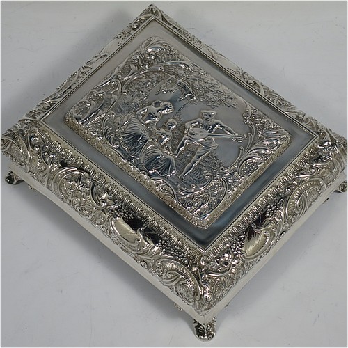 A very pretty Antique Victorian Sterling Silver casket style jewellery dressing table box, having a rectangular shaped casket style body, with hand-chased floral and scroll decoration, a hinged lid with a hand-chased figural scene, a dark green velvet-lined interior, and all sitting on four shell feet. Made by Henry Matthews of Birmingham in 1900. The dimensions of this fine hand-made antique silver dressing table jewellery box are length 16 cms (6.25 inches), width 13.5 cms (5.25 inches), and height 7 cms (2.75 inches).