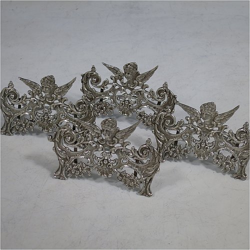 An Antique Sterling Silver set of four place card holders, having cast bodies with winged seraphims over scroll and floral decoration, and sitting on half-circle wire-work bases. Made by Stuart Clifford of London in 1904. The dimensions of this fine set of antique silver place card holders are width 7.5 cms (3 inches), height 5 cms (2 inches), depth 4 cms (1.5 inches), and they weigh a total of approx. 101g (3.3 troy ounces).