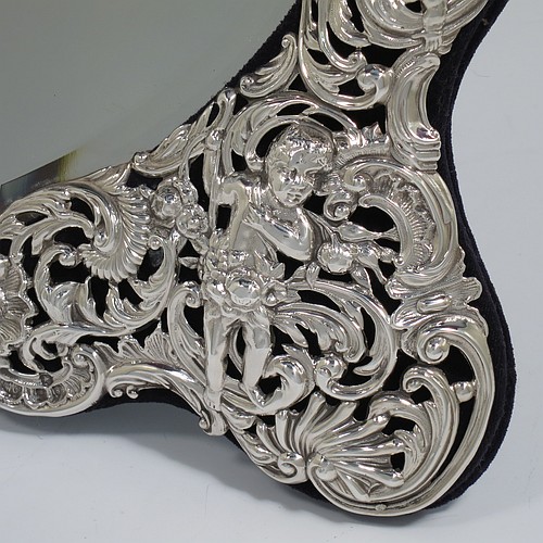An Antique Victorian Sterling Silver table or vanity mirror having hand-chased cherubs and birds, scroll-work, and floral decoration, with a top vacant cartouche, an original bevelled heart-shaped mirror, and blue velvet backing with easel stand. Made by William Comyns of London in 1893. The dimensions across the frame of this fine hand-made antique silver vanity or table mirror are height 28 cms (11 inches), and width 24 cms (9.5 inches).
