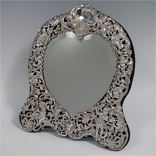An Antique Victorian Sterling Silver table or vanity mirror having hand-chased cherubs and birds, scroll-work, and floral decoration, with a top vacant cartouche, an original bevelled heart-shaped mirror, and blue velvet backing with easel stand. Made by William Comyns of London in 1893. The dimensions across the frame of this fine hand-made antique silver vanity or table mirror are height 28 cms (11 inches), and width 24 cms (9.5 inches).