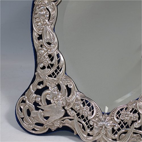 An Antique Victorian Sterling Silver table or vanity mirror having hand-chased birds, scroll-work, and floral decoration, with a top vacant cartouche, an original bevelled heart-shaped mirror, and blue velvet backing with easel stand. Made by William Comyns of London in 1897. The dimensions across the frame of this fine hand-made antique silver vanity or table mirror are height 28 cms (11 inches), and width 24 cms (9.5 inches).