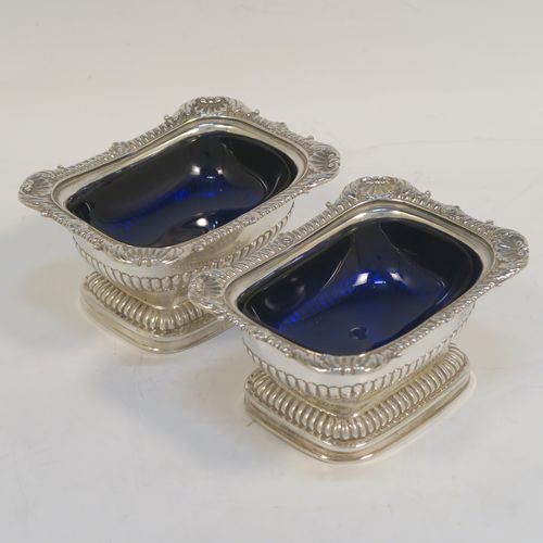 A large and handsome pair of Antique Georgian Sterling Silver salt cellars in a Regency style, having rectangular bodies, with blue-glass liners, applied shell and gadroon borders, half-fluted hand-chased decoration, and all sitting on pedestal bases. These elegant antique silver salt cellars were made in London in 1811. The dimensions of these fine hand-made antique silver salt cellars are length 9.5 cms (3.75 inches), width 7 cms (2.75 inches), height 5 cms (2 inches), and they weigh a total of approx. 237g (7.6 troy ounces). Please note that these items are part crested.   