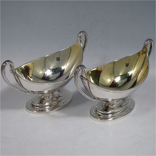 An Antique Georgian Sterling Silver pair of salt cellars, in a boat-shaped neoclassical style, having plain oval bodies, with gold-gilt interiors, applied reeded borders, and reeded side-handles, all sitting on pedestal feet. Made by John Wakelin and William Taylor of London in 1785 (having the rare Incuse duty mark). The dimensions of these fine hand-made antique silver salt cellars are length 13.5 cms (5.3 inches), width 6.5 cms (2.5 inches), height 8 cms (3.25 inches), and they weigh a total of approx. 214g (6.9 troy ounces). Please note that both salt cellars have engraved crests.