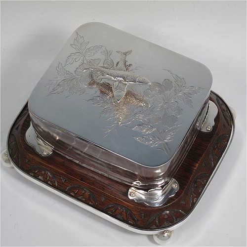 A very handsome Antique Victorian Silver Plated sardine dish, having a rectangular body with rounded corners, a cut-crystal liner sitting in a frame attached to an Oak wooden base with four ball feet, a lift-off lid with cast crossed sardines above hand-engraved floral decoration. Made in ca. 1890. The dimensions of this fine hand-made antique silver-plated sardine dish are length 18 cms (7 inches), width 15 cms (6 inches), and height 10 cms (4 inches).