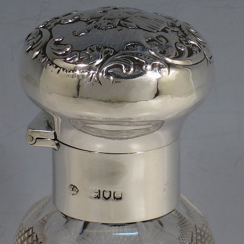 An Antique Victorian Sterling Silver and hand-cut crystal large table scent bottle, having a hand-chased round hinged lid with floral and figural decoration, a plain round mount, an internal stopper, and an unusual shaped hand-cut crystal body with floral, swags, bows, and fluted decoration. Made by William Comyns of London in 1900. The dimensions of this fine hand-made antique silver & crystal scent bottle are height 18 cms (7 inches), and diameter 10 cms (4 inches).    