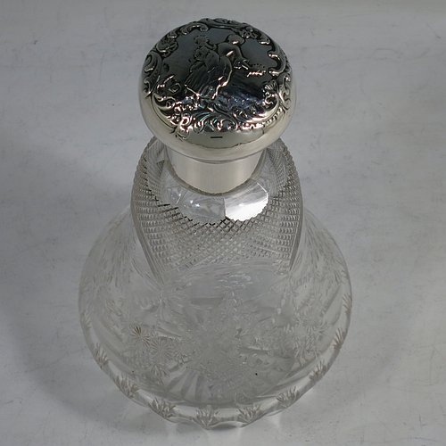 An Antique Victorian Sterling Silver and hand-cut crystal large table scent bottle, having a hand-chased round hinged lid with floral and figural decoration, a plain round mount, an internal stopper, and an unusual shaped hand-cut crystal body with floral, swags, bows, and fluted decoration. Made by William Comyns of London in 1900. The dimensions of this fine hand-made antique silver & crystal scent bottle are height 18 cms (7 inches), and diameter 10 cms (4 inches).    