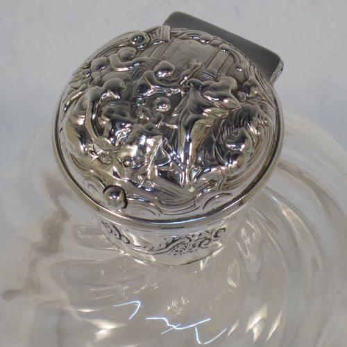 A very pretty and unusual pair of Antique Edwardian Sterling Silver and hand-cut crystal table scent bottles, having round mounts with hinged lids and internal stoppers, the lids with hand-chased figural scene decoration and push-button snap fits. The crystal bodies with rounded square cross-sections and swirl fluted decoration, and all sitting on flat bases. Made by William Comyns of London in 1907. The dimensions of these fine hand-made antique silver and crystal scent bottles are height 12.5 cms (5 inches), and 8.5cms (3.3 inches) square.  
