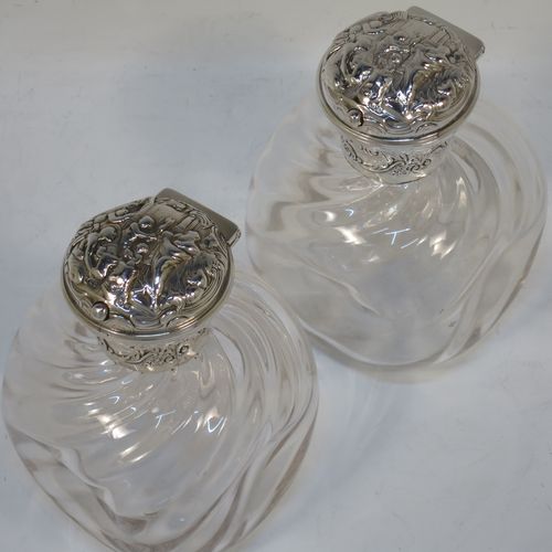 A very pretty and unusual pair of Antique Edwardian Sterling Silver and hand-cut crystal table scent bottles, having round mounts with hinged lids and internal stoppers, the lids with hand-chased figural scene decoration and push-button snap fits. The crystal bodies with rounded square cross-sections and swirl fluted decoration, and all sitting on flat bases. Made by William Comyns of London in 1907. The dimensions of these fine hand-made antique silver and crystal scent bottles are height 12.5 cms (5 inches), and 8.5cms (3.3 inches) square.  