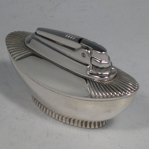 A Sterling silver table lighter, having a plain oval lower body, with fluted shoulders, and sitting on a fluted collet foot. The lighter mechanism is by Ronson and is in fully working order. Length 11 cms (4.25 inches), height 6 cms (2.3 inches), width 4.5 cms (1.75 inches). Please note that this item is crested.
