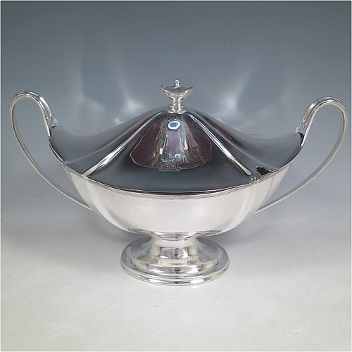 An Antique Edwardian Sterling silver Neoclassical style soup tureen, having a pull-off lid with an urn-shaped finial and cut-out slot for a ladle, with hand-chased shoulders, applied reeded borders, two reeded scroll side-handles, and all sitting on a pedestal foot. Made by Goldsmiths & Silversmiths  of London in 1902. The dimensions of this fine hand-made antique silver soup tureen and cover are height 23 cms (9 inches), length including handles 38 cms (15 inches), width 20 cms (8 inches), and it weighs approx. 1,615 (52 troy ounces). Please note that this item is crested on lid and body.