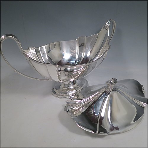 An Antique Edwardian Sterling silver Neoclassical style soup tureen, having a pull-off lid with an urn-shaped finial and cut-out slot for a ladle, with hand-chased shoulders, applied reeded borders, two reeded scroll side-handles, and all sitting on a pedestal foot. Made by Goldsmiths & Silversmiths of London in 1902. The dimensions of this fine hand-made antique silver soup tureen and cover are height 23 cms (9 inches), length including handles 38 cms (15 inches), width 20 cms (8 inches), and it weighs approx. 1,615 (52 troy ounces). Please note that this item is crested on lid and body.